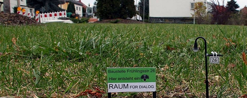 Schild auf Rasen: Baustelle Frühlingsstr. 26. Hier entsteht ein Raum für Dialog. Von Grass Lifter.
