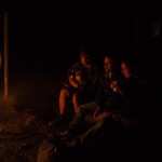 Drei Personen trinken Bier am Lagerfeuer bei Nacht