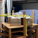 Kunstobjekt: Mehrere Tische aufeinander gestapelt mit eingepackten Paketen und Blättern oben drauf, mit einem "Attention" Band um die Beine des obersten Tischs.