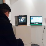 Person schaut die Podiumsdiskussion auf einem kleinen Röhrenfernseher und schaut gleichzeitig daneben auf einen Laptop, auf dem Musik abgespielt wird.