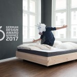 Person im Hemd und mit ausgebreiteten Armen und Einhornkopf auf einem Bett mit Logo German Design Award Nominee 2017 (Foto von Julia Kneuse)