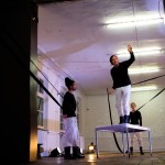 Ein Schauspieler steht auf einem Tisch, zwei Schauspielerinnen bzw. Schauspieler schauen ihn an.