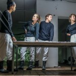 Schauspielende stehen vor einem Tisch, zwei sind durch Bänder verbunden und eine andere Person schaut weg aus dem Bildausschnitt, wohin ihr Band geht.