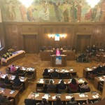 Bild von oben auf ein Pult, an dem eine Rede vor sitzenden Menschen gehalten wird.