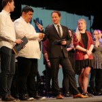 Ein Schüler hält eine Rede neben einem Moderator und den anderen Schülerinnen und Schülern auf der Bühne