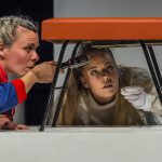 Zwei Schauspielerinnen beim Stück "Aufstand der Dinge" untersuchen eine Sitzbank von unten, die auf einem Tisch steht und an deren Boden etwas befestigt ist