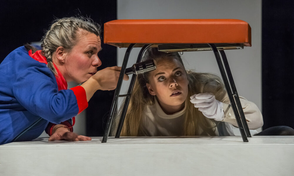 Zwei Schauspielerinnen beim Stück "Aufstand der Dinge" untersuchen eine Sitzbank von unten, die auf einem Tisch steht und an deren Boden etwas befestigt ist