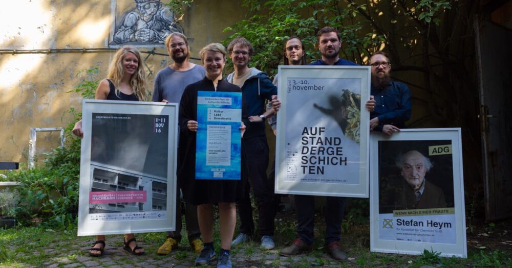 Preistragende des Kultur.LEBT.Demokratie Preises vom Asa-FF stehen mit Urkunde und drei gerahmten Plakaten der jährlichen Theaterfestivals zwischen 2016 und 2018 im Innenhof des Kabinettstückchens am Fuße des Sonnenbergs