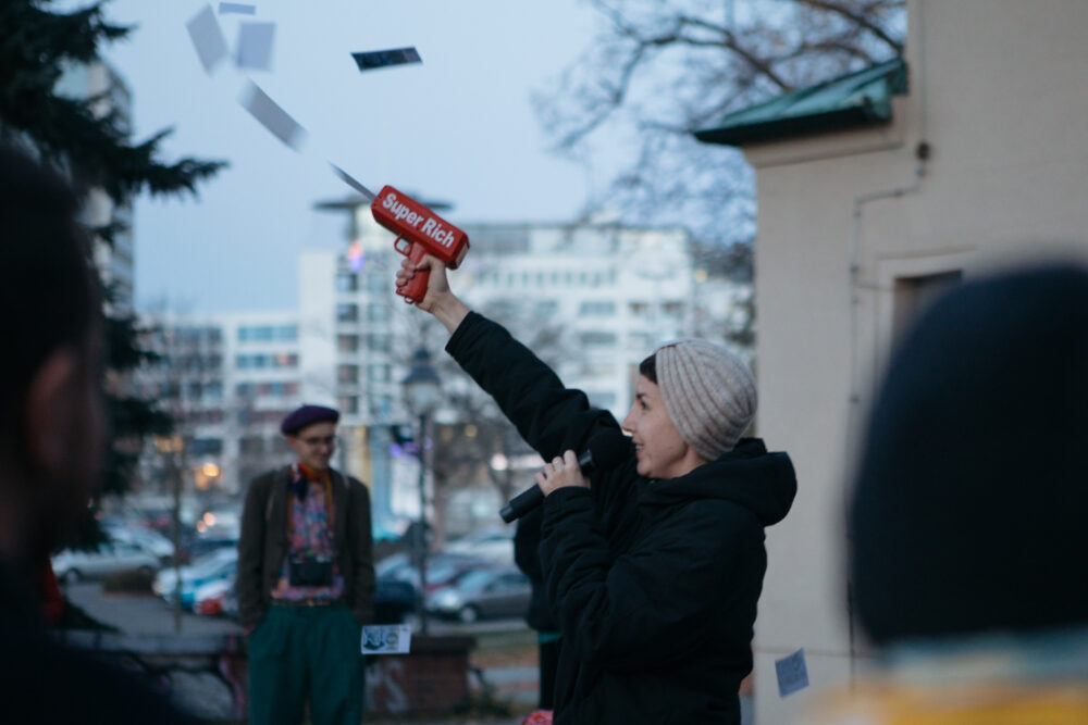Schüsse in die Luft Aktivistin feuert falsche Geldscheine mit einer Pistole draußen in die Luft, auf der "Super Rich" steht und lacht während sie das Mikrofon vors Gesicht hält