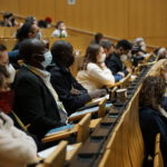 Menschen sitzen in einem Hörsaal, sie sehen nach vorn und tragen Mund-Nasen-Schutz