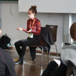 Eine Frau hält einen Vortrag. Eine Gruppe von Menschen hört ihr zu.