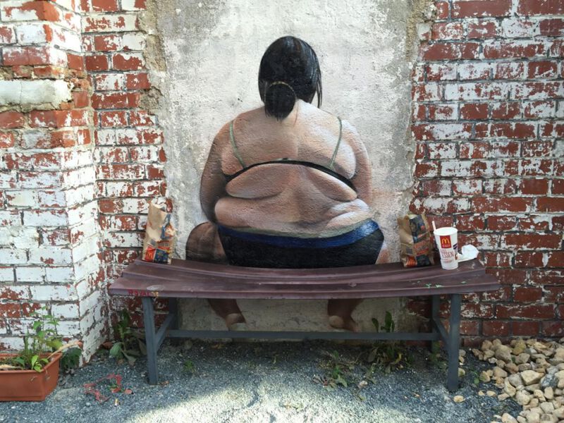 Mauergrafitti übergewichtige Frau in Unterwäsche von hinten. Vor der Mauer steht eine Bank, auf der links und rechts neben dem Motiv McDonaldsverpackungen liegen.