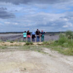 Am Grubenrand Aktivistinnen und Aktivisten stehen in der Ferne und sind von hinten zu sehen vor dem Braunkohletagebau. Ein Mitglied einer lokalen Bürgerinitiative erklärt die Lage.