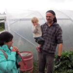 Besuch bei der CSA Sehlis Ein Aktivist mit Kleinkind im Arm lächelt in Richtung einer Frau, die gerade in einen Rhabarberstängel beißt.