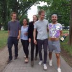 Fünf lachende Menschen gehen durch den Park. Davon ein Mann im bauchfreien Glitzershirt und Sektflasche in der Hand.