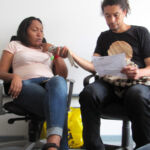 Im Interview Juan Manuel Ortega beim Interview: Zwei People of Color sitzen in einem Stuhl, eine Person hält der anderen ein Smartphone als Aufnahmegerät hin.