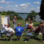 Teilnehmende beim Besprechen im Stuhlkreis in der Sonne mit See im Hintergrund