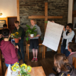 Zwei Teilnehmer präsentieren den anderen Teilnehmenden etwas mit Blumentöpfen in der Hand, während ein anderer Teilnehmer ein Plakat hochhält.