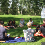 Teilnehmende beim Impulse Boost Camp sitzen im Kreis auf einer Wiese im Freien, während der Asa Referent Franz Knoppe etwas an einer Plakatwand präsentiert