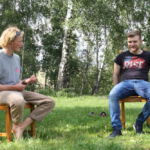 Zwei Teilnehmende sitzen auf Holzstühlen im Freien auf der Wiese mit etwas Abstand, während die eine Person in Richtung der anderen Person spricht