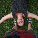 Eine glückliche Frau liegt mit ausgebreiteten Armen auf einer grünen Wiese zwischen den Füßen einer Person, die lackierte Fußnägel hat und Flip Flops trägt.