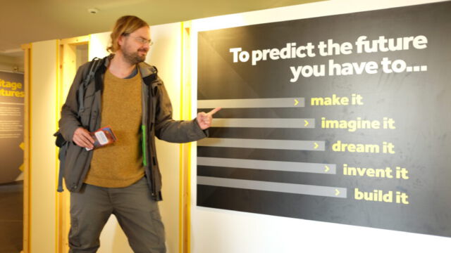Ausstellung Heritage Futures im Manchester Museum (c)Frauke Frech Franz vom Asa-FF zeigt grinsend auf ein Wandbild der Heritage Futures Ausstellung im Manchester Museum: "To predict the future you havve to... make it, imagine it, dream it, invent it, build it" steht darauf.