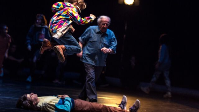 INVITED Bild einer großen Performance im Rahmen vom Aufstand der Utopien: Ein Junge ist beim Sprung in der Luft mit angewinkelten Beinen zu sehen, während er sich über einem Teenager befindet. Dieser Teenager liegt mit geschlossenen Augen am Boden. Im Licht eilt ein älterer Herr heran, andere Schauspielerinnen gehen im Dunkeln weiter entfernt um den Teenager kreisend herum