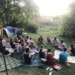 Teilnehmende des Sommerlagers sitzen entspannt auf der Wiese auf Decken. Manche trinken Bier. Zwei aufgestellte Zelte sind zu sehen.