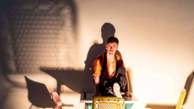 Ein Schauspieler sitzt mit einem ausgestreckten Bein und einem angewinkeltem Bein auf dem Tisch und schaut ernst in Richtung Kamera