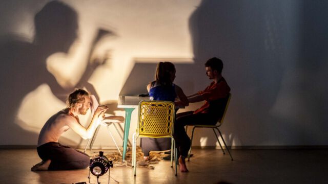 Konzept Europa, Rico Wagner, Elisabeth Caesar, Jule Heidelbach, Marie Lemser, Max Howitz, Martin Schnippa; Theater an der Parkaue, Park an der Parkaue, Ballhaus Ost Oberkörperfreier Schauspieler kniet auf dem Boden und macht eine Geste mit den Händen, während die zwei Schauspielende am Küchentisch sitzen und ihn nicht beachten