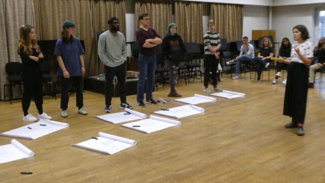 Schülerinnen und Schüler stehen in einer Reihe vor weißen Plakaten. Dabei liegt an jedem Plakat ein Edding für die Beschriftung.