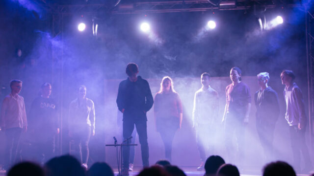 Schauspielende auff der Bühne starren in Richtung eines Schauspielers im Mittelpunkt. Es ist recht dunkel und nur ein paar Scheinwerfer sind an. Der Nebel aus einer Nebelmaschine ist klar erkennbar.