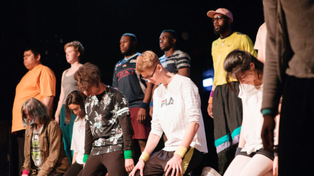 Eine Reihe von Schauspielenden steht vor einer Reihe von knienden Schauspielenden, die auf den Boden starren