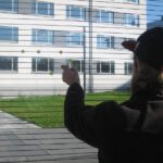 Random Music. Foto© Utopia Toolbox Ein Mann schaut aus dem Fenster. Auf diesem sind gelbe Punkte aufgeklebt, nach denen er greift. Im Hintergrund ist das Gebäude des Studentenwerks am Thüringer Weg zu sehen