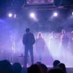 Szene vom Wir. Wie? Theaterstück in Zwickau: Ein Schauspieler steht im Mittelpunkt und schaut auf den Boden. Acht andere Schauspielende stehen im Halbkreis um ihn und schauen ihn an. Durch die Beleuchtung lässt sich der Nebel aus der Nebelmaschine deutlich erkennen